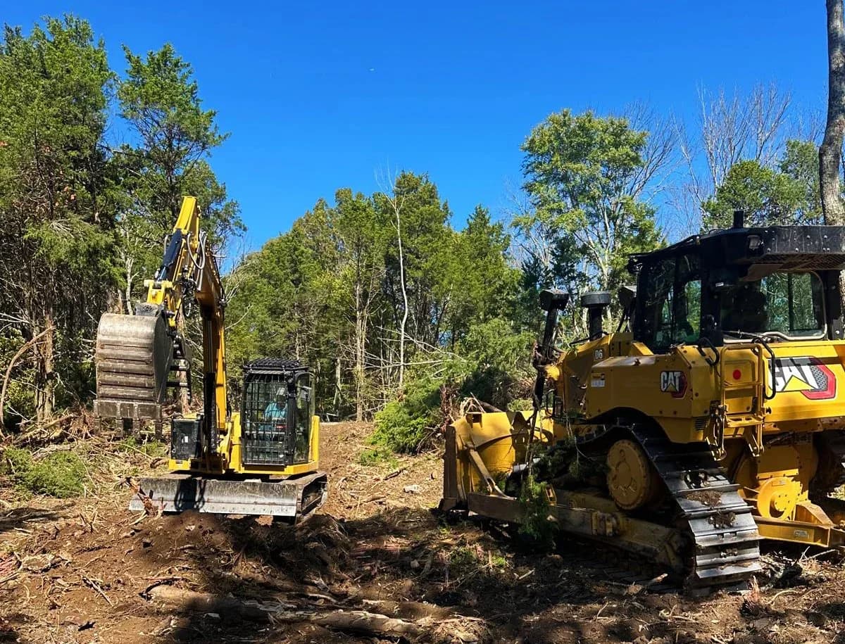 Two clean CAT machines working a wooded clearing site against a bright Tennessee sky.